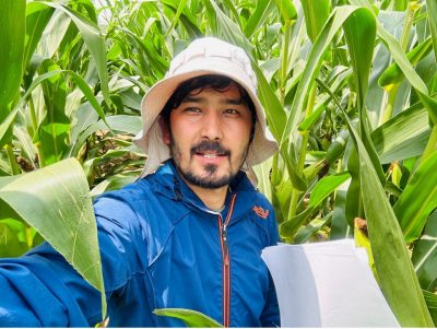 A young man wearing a bucket hat stands in a corn field holding papers